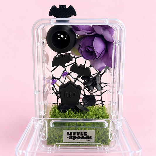 Open front view of a gothic juvenile jumping spider enclosure with purple flowers, black bat decorations, gravestone, and moss